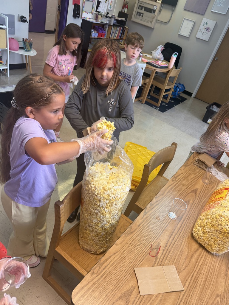 Students wearing gloves scoop popcorn from a large bag while preparing individual snack bags in a classroom.