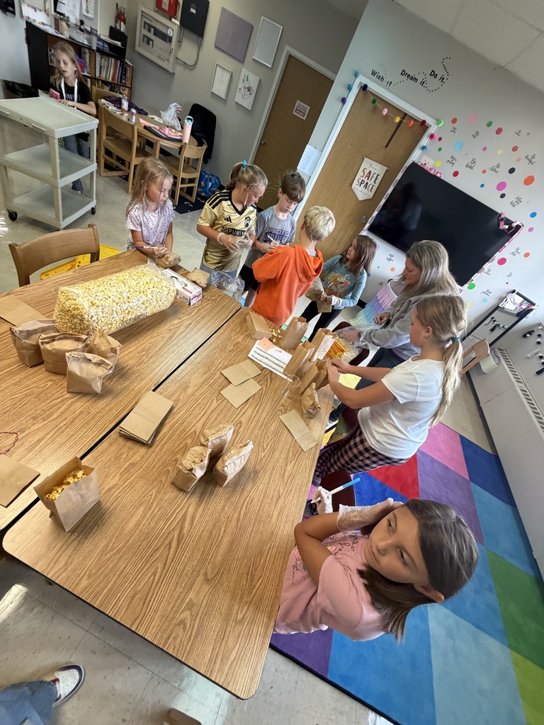 Students and a teacher work together around classroom tables to fill brown paper bags with popcorn.