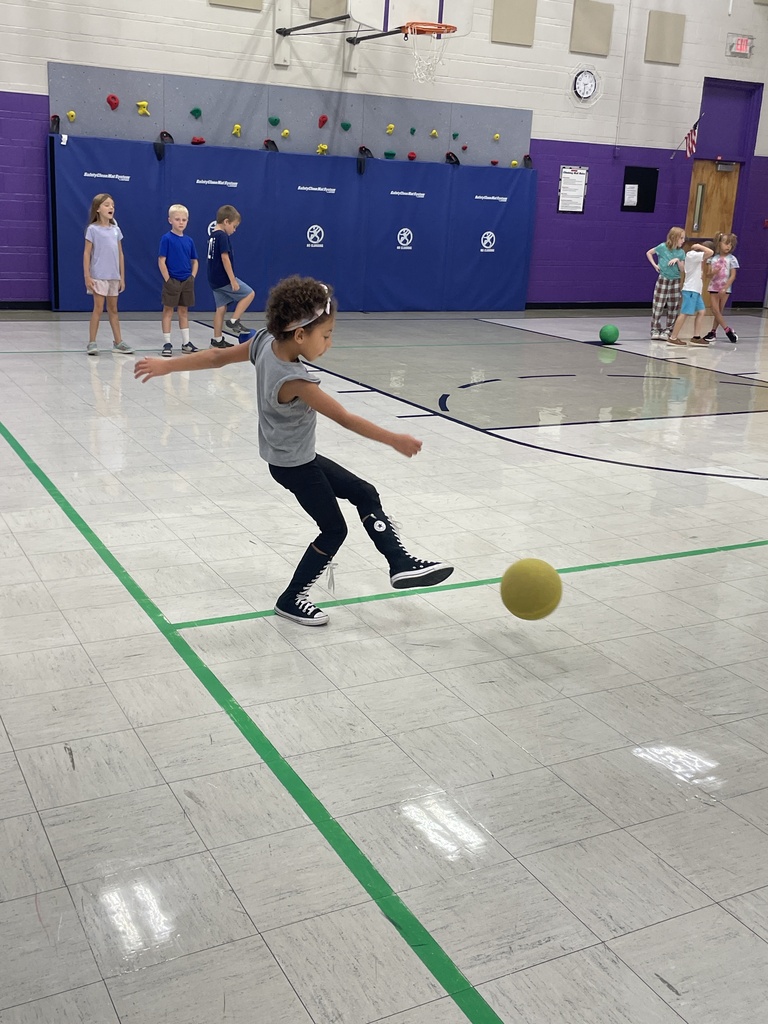 A student kicks a yellow ball across the gym floor while classmates wait nearby during a physical education activity.