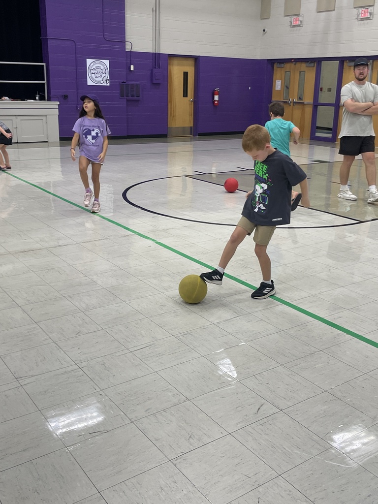 A student stops a yellow ball with his foot as classmates participate in a gym activity and a teacher observes.