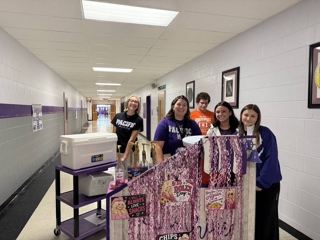 Students and staff smile beside a decorated snack cart with coolers and flavored drink syrups in a school hallway.