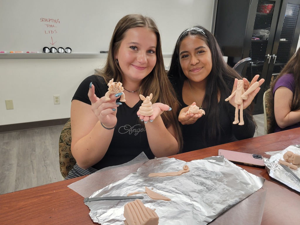 Two students smile and display small handmade clay figures they sculpted during an art class.