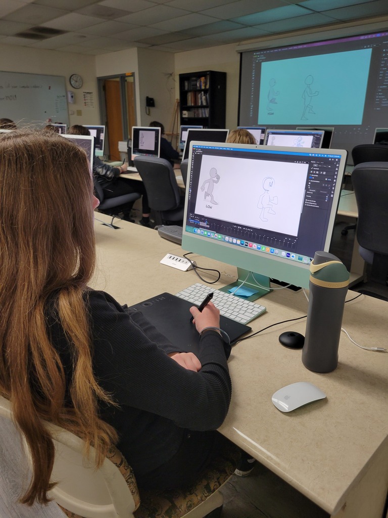 Student uses a drawing tablet and computer to create a character animation while classmates work at nearby stations.