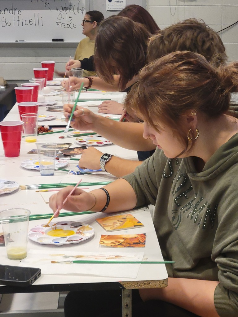 Students focus on painting small artworks at a classroom table covered with brushes, palettes, cups, and reference images.