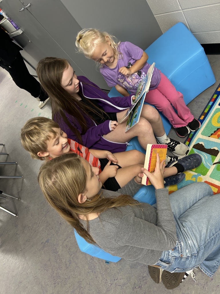 Two older students share books with two younger children while seated together on bright blue classroom chairs.
