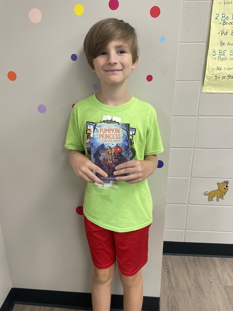 Student smiles while holding the book “The Pumpkin Princess and the Forever Night” in a colorful classroom.