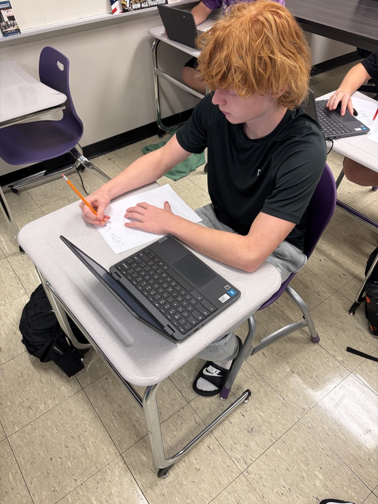 Student works at a classroom desk, writing on paper beside an open laptop during an assignment.