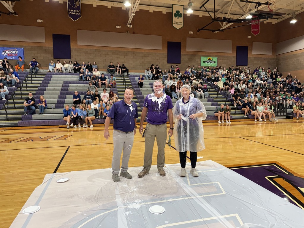 Three school staff members covered in whipped cream smile on the gym floor as students watch from the bleachers.