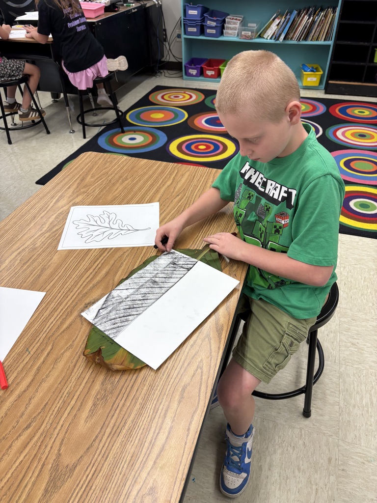 A student uses a black crayon to make a leaf rubbing on white paper during a classroom art project.
