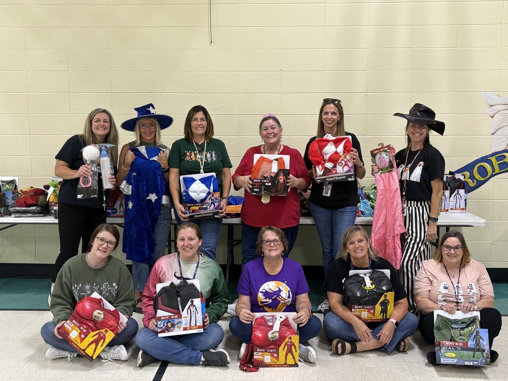 Ten smiling school staff members pose in two rows, holding Halloween costumes, including superheroes and princess outfits.