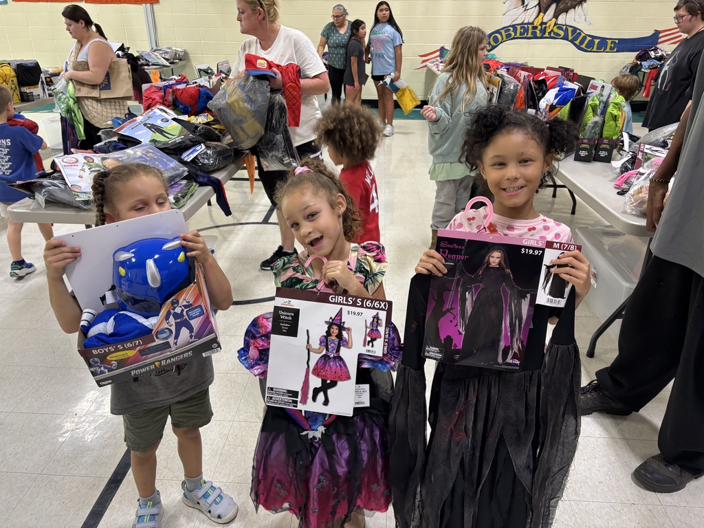 Three smiling children hold up Power Rangers, unicorn witch, and Soul Reaper Halloween costumes at a school costume event.