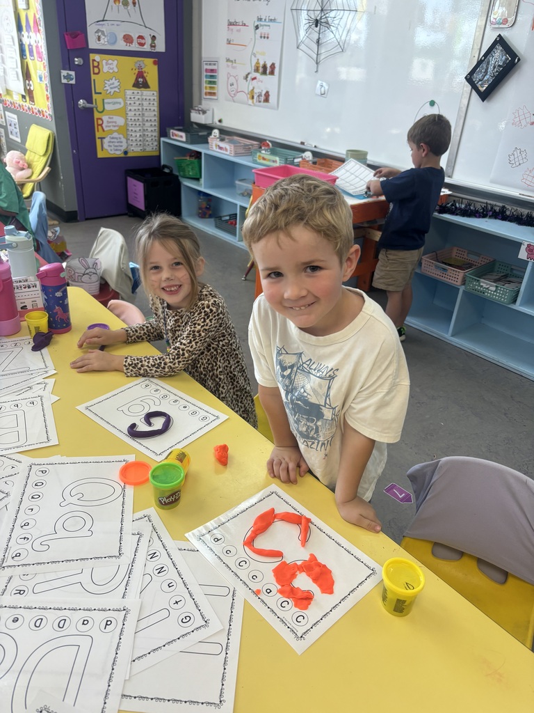 Two smiling students create letters with colorful Play-Doh on alphabet worksheets at a classroom activity table.