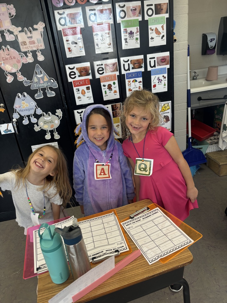 Three smiling elementary students wearing letter necklaces pose beside a desk with worksheets and classroom supplies.