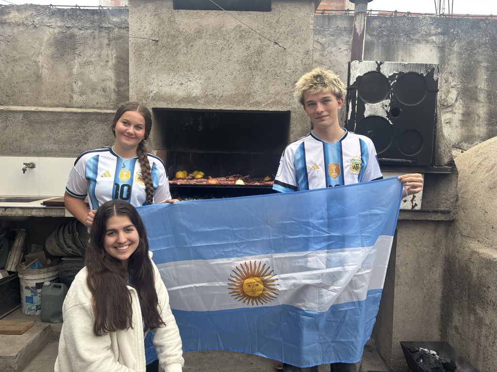 Three smiling students pose with an Argentine flag in front of an outdoor grill, two wearing Argentina soccer jerseys.