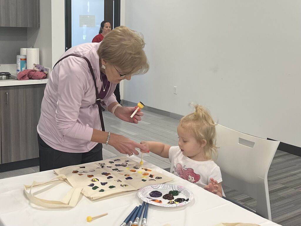 A woman helps a young girl use colorful paint and sponge brushes to decorate a canvas tote bag.