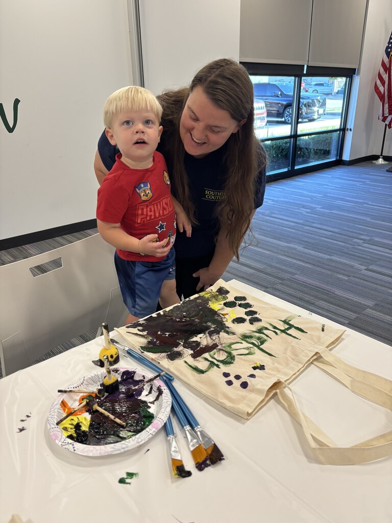 A smiling woman stands beside a young boy displaying his painted tote bag covered in colorful designs.