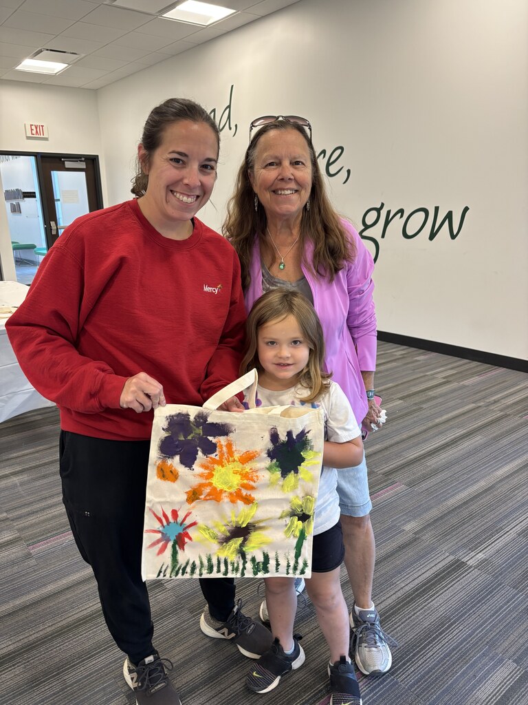 Two smiling women and a young girl proudly display a tote bag decorated with bright, colorful painted flowers.