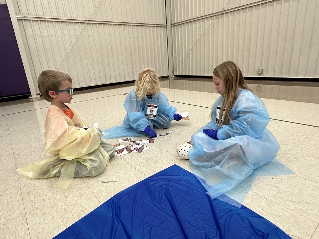 Three students wearing medical gowns and gloves sit on the gym floor, working together on a hands-on educational activity.