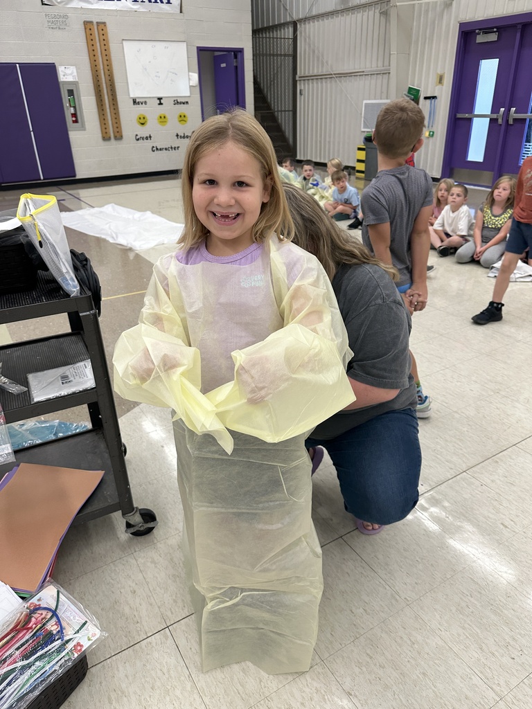 A smiling young girl models a yellow medical gown while an adult helps her prepare for a classroom medical learning activity.