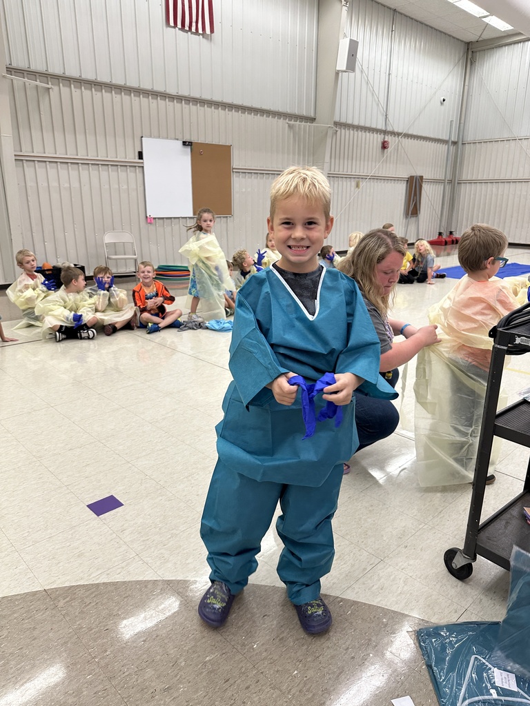 A smiling young boy wears teal medical scrubs and holds blue gloves while classmates prepare for a hands-on learning activity.