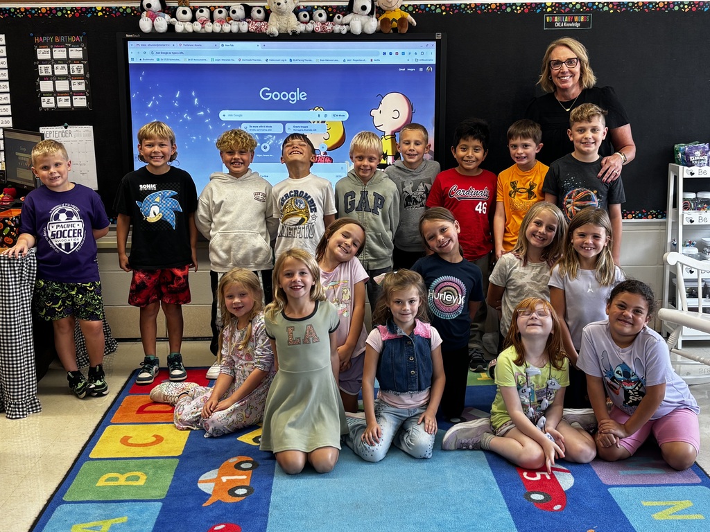 A smiling teacher poses with a group of elementary students on a colorful classroom rug in front of a digital display.