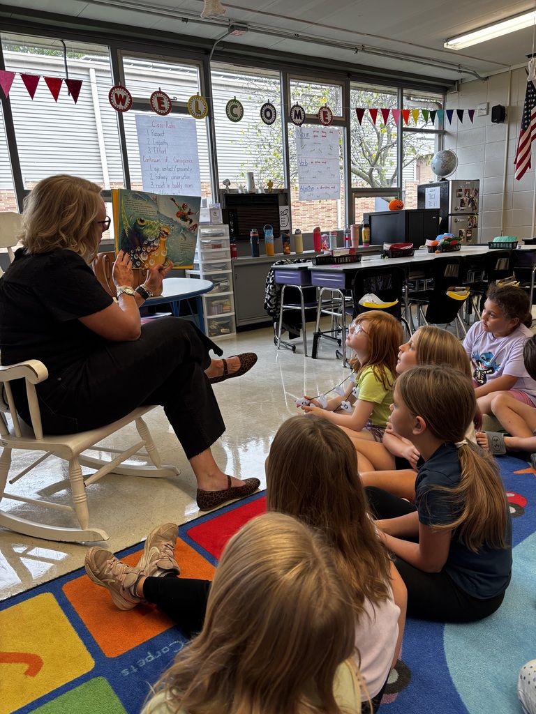 A woman reads a colorful picture book aloud to elementary students seated on a classroom rug, listening attentively.