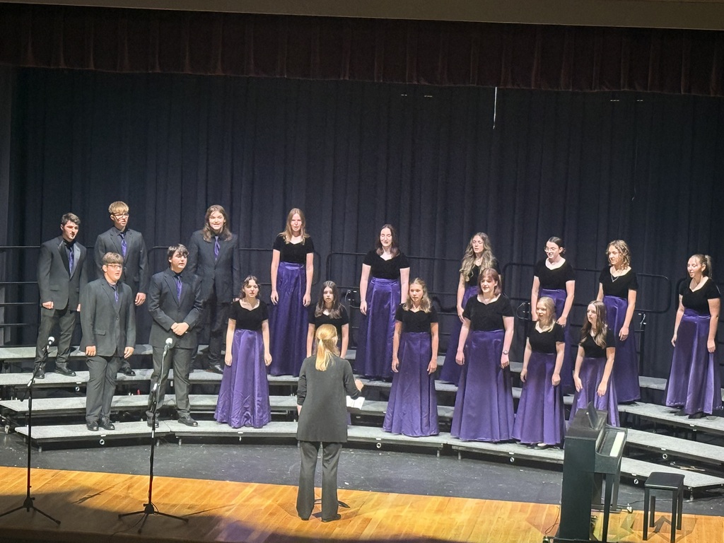 A student choir performs on stage in black and purple formal attire, guided by a conductor standing in front of the group.