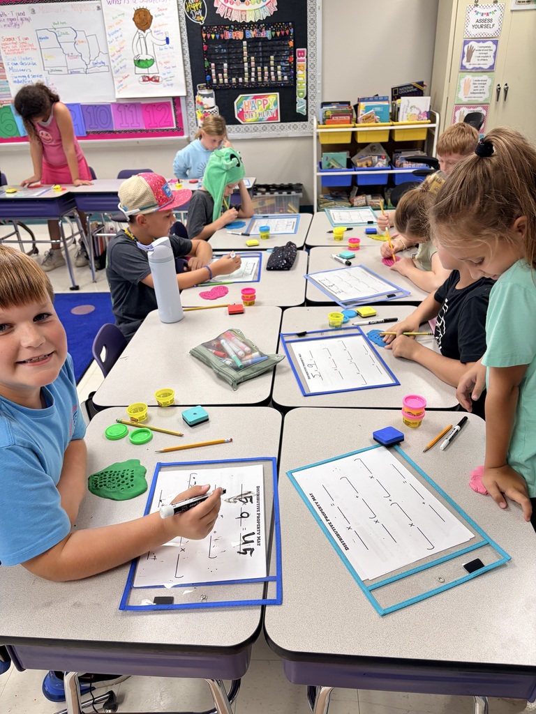 Students work on multiplication worksheets using colorful modeling clay, pencils, and markers during a hands-on math lesson.