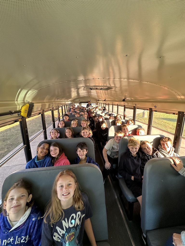 Smiling students fill the seats of a school bus, looking toward the camera in warm afternoon sunlight.