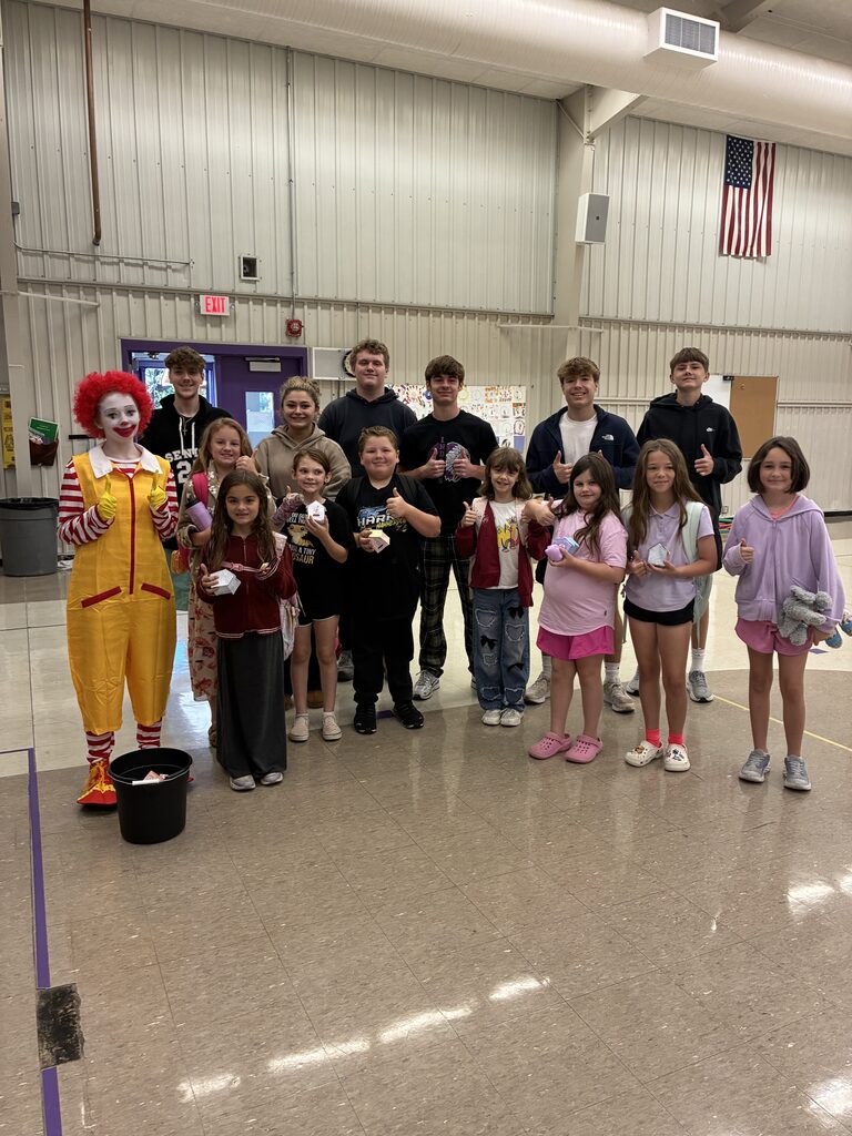 Students of various ages pose with a student dressed as Ronald McDonald in a school gymnasium.