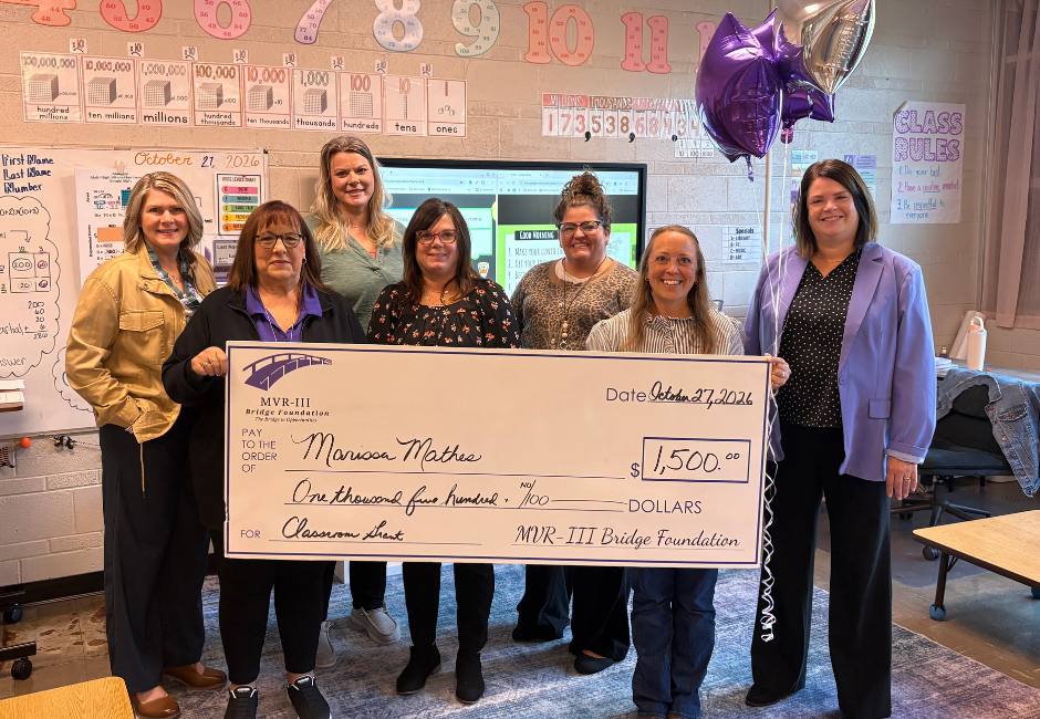 Various staff members pose with a large check in a school classroom.