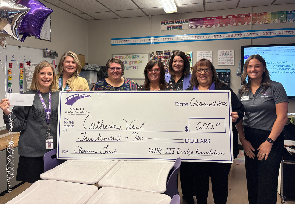 Various staff members pose with a large check in a school classroom.