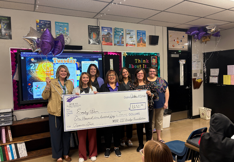 Various staff members holding a large check in a school classroom.
