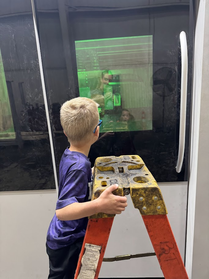 A child stands on a ladder, looking at a green screen displayed on a window, possibly in a classroom.