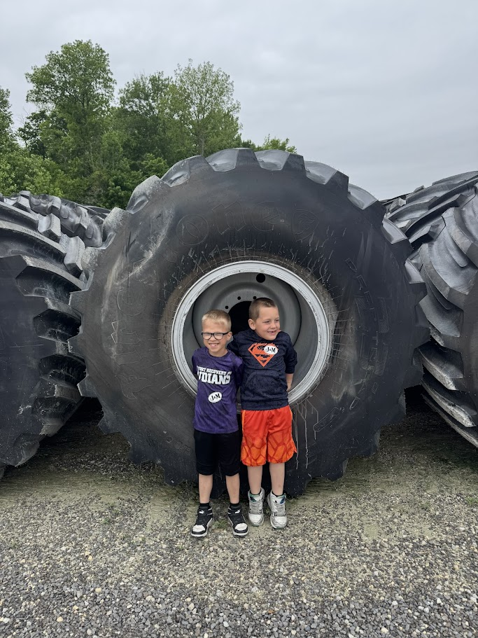 Two children pose in front of a large vehicle tire. They stand on a gravel surface with trees in the background.