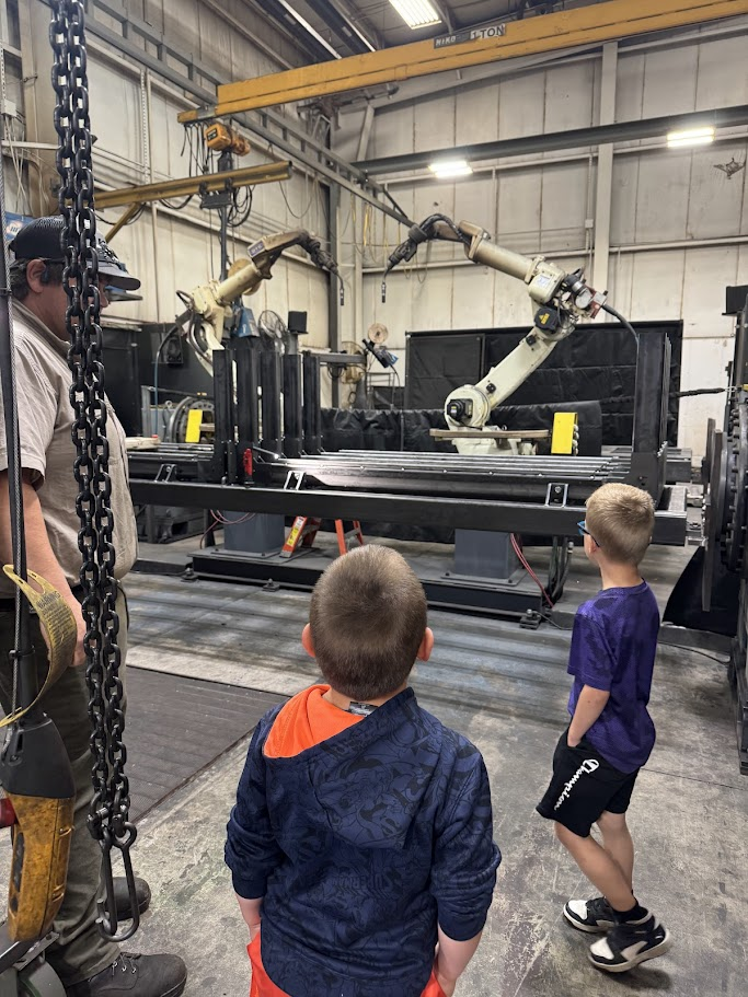 Factory setting with two young boys observing an adult operating machinery. A robotic arm is active.