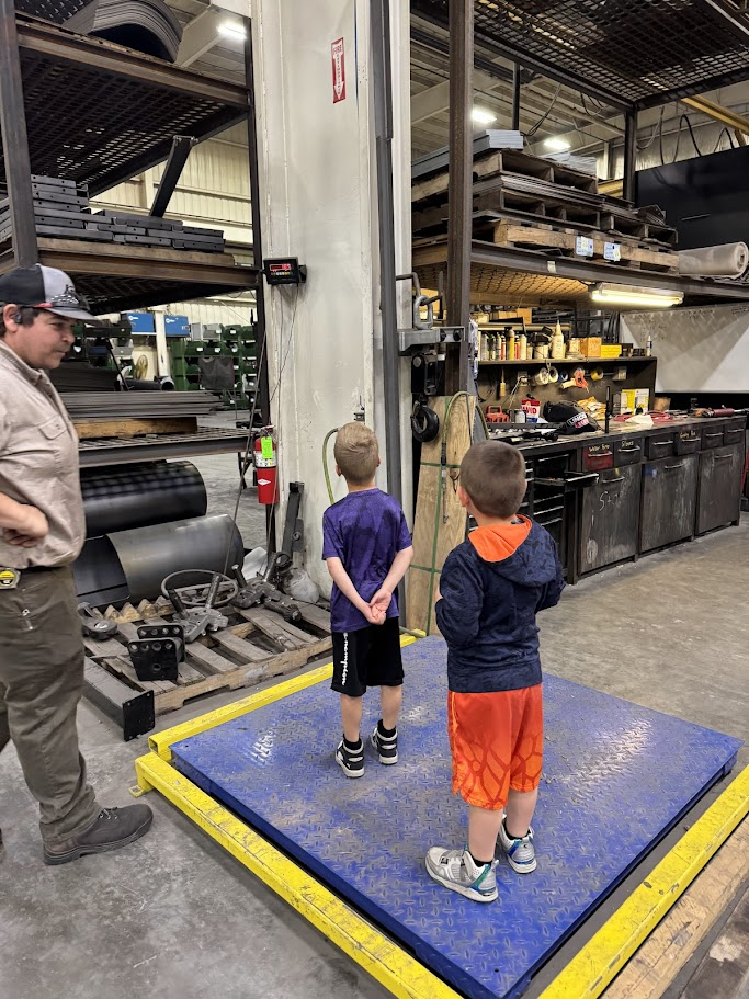 A man instructs two boys on a blue platform in a workshop. They are standing on a blue mat. Shelves, tools, and equipment surround them.