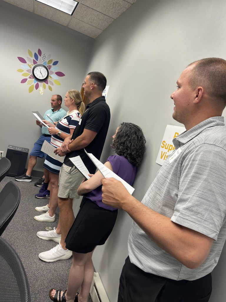 Group of people standing in a room, reading from a paper. Wall with clock decoration.