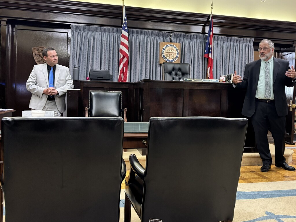 Two men in suits, one speaking, stand in a formal room with leather chairs, a long desk, and flags behind.