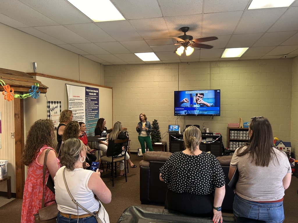 People in a room with a television on the wall and a woman standing in front of a small crowd.