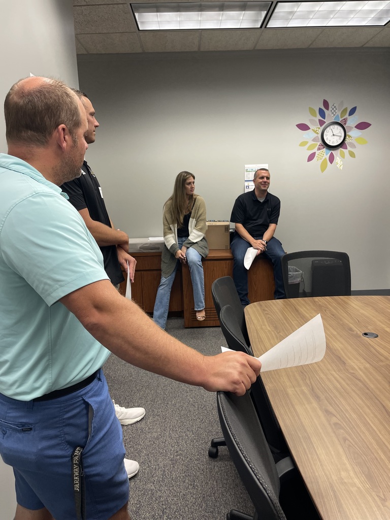 People in a meeting room. A man stands, holding a paper. A woman sits on a bench. Another man sits on a chair. A clock is on the wall.