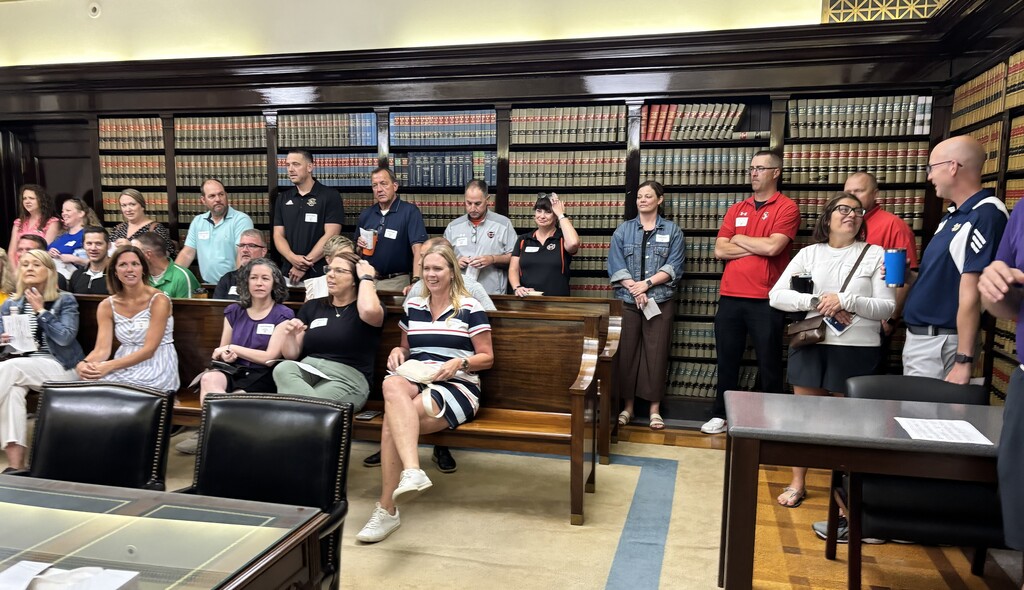 Group of people in a room with wooden benches, bookshelves, and tables; some are seated, others are standing.