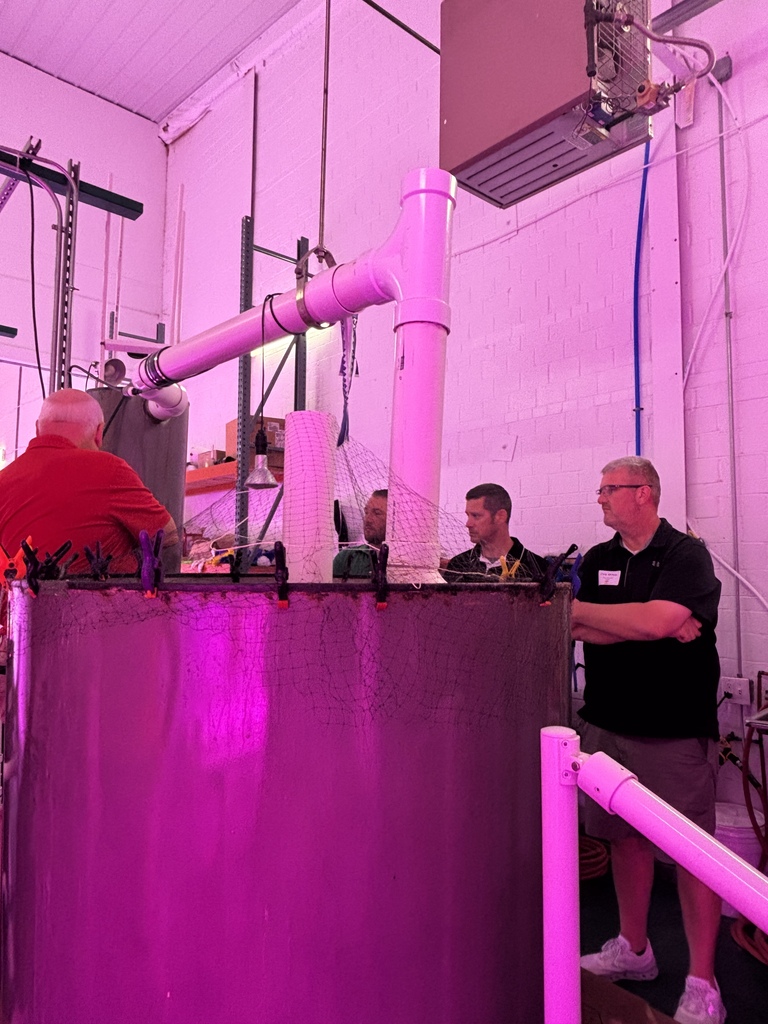 Three men in a room with pink lighting. The men are looking at a large, cylindrical container.