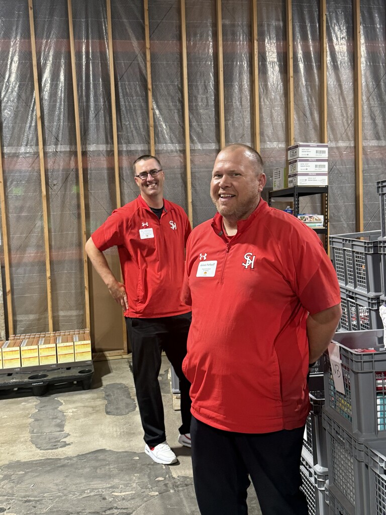 Two men in red polo shirts and black pants stand in front of plastic crates and a white curtain.