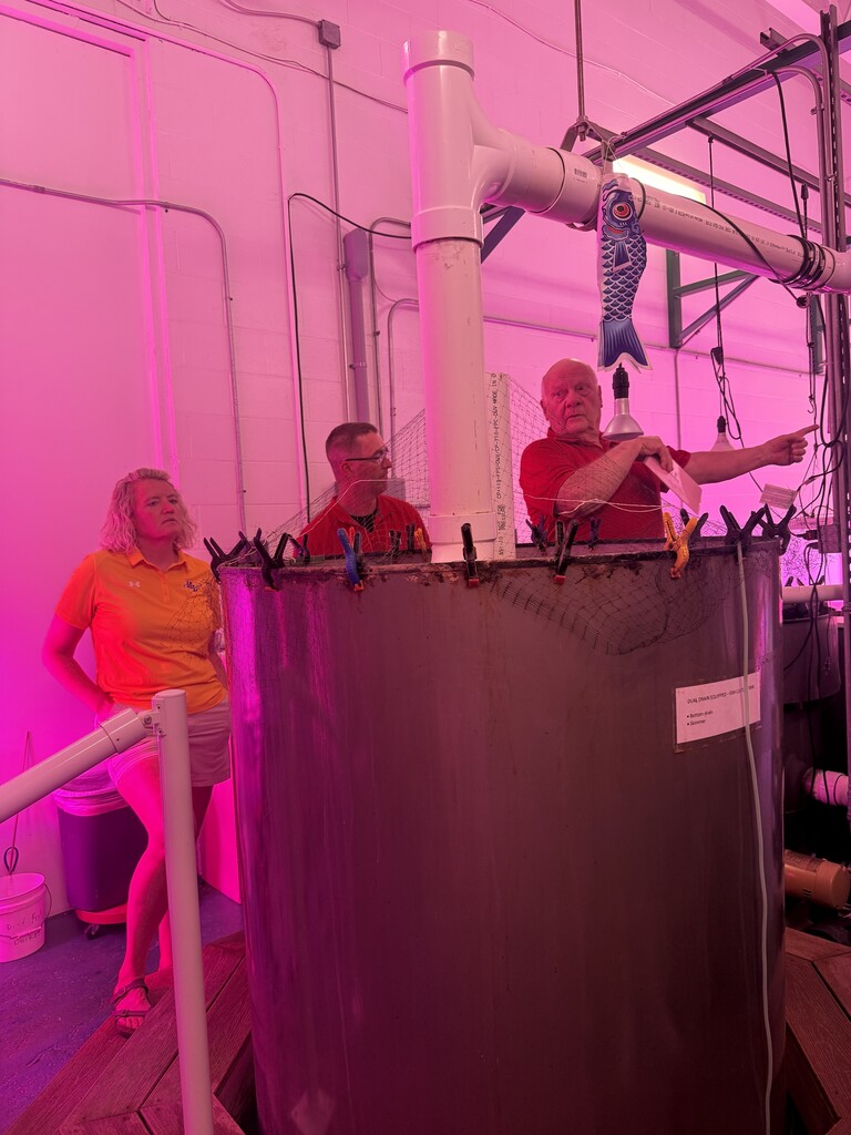 Three people stand around a large metal tank with a white pipe. A woman and a man look at the tank.