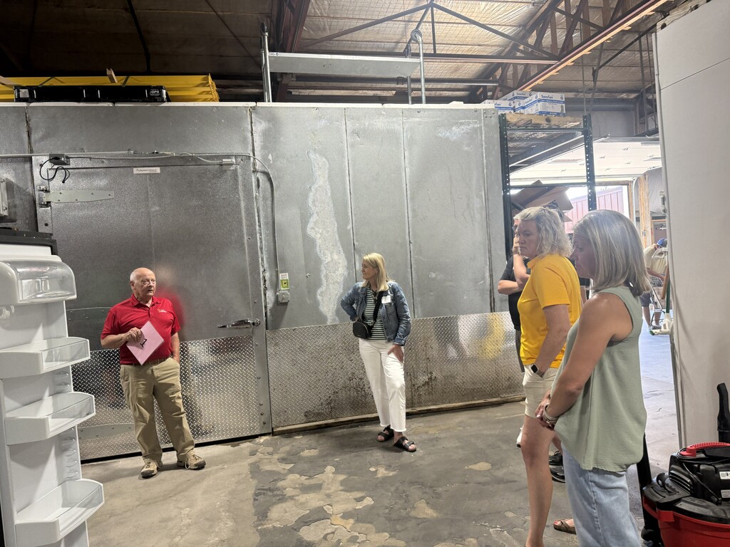 A group of people inside a warehouse, one man in red shirt holding paper, stands near a large metal door.