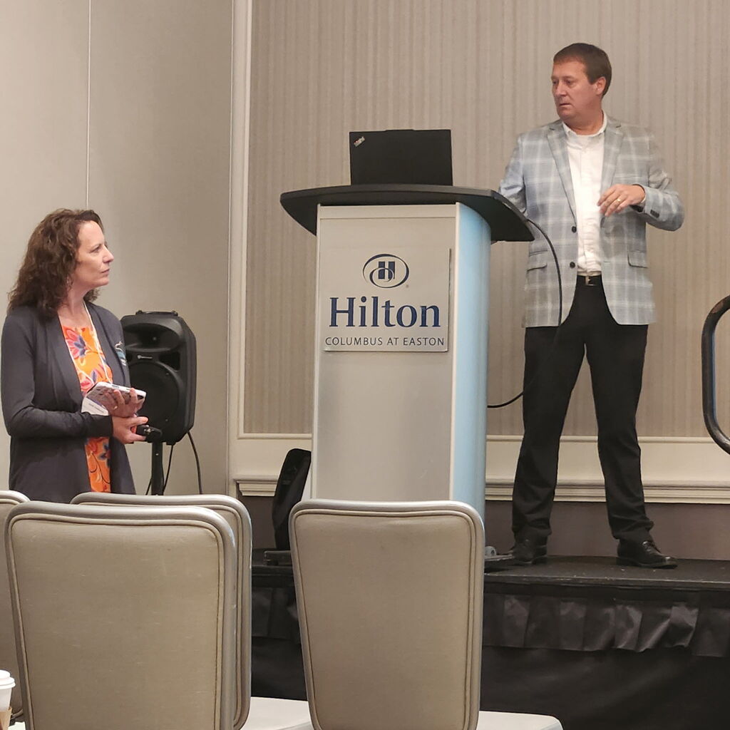 Man and woman standing by a podium with a laptop on it. Behind them is a Hilton sign.