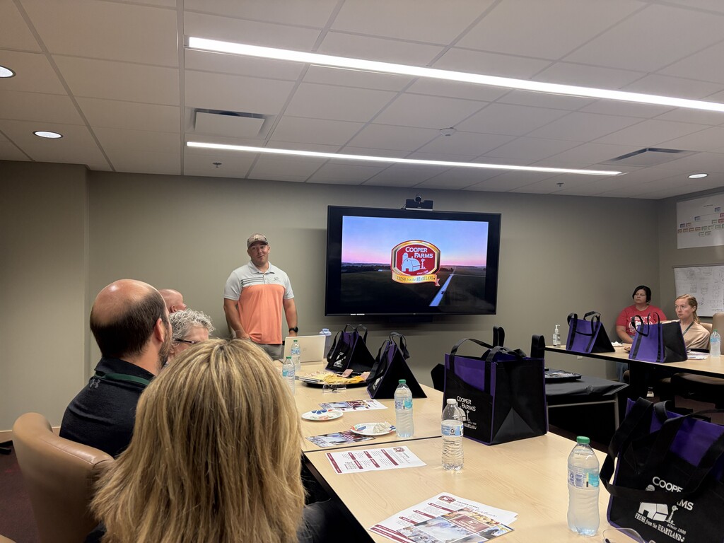 A person gives a presentation in a room. A large screen displays behind them. A table holds bottles and bags.