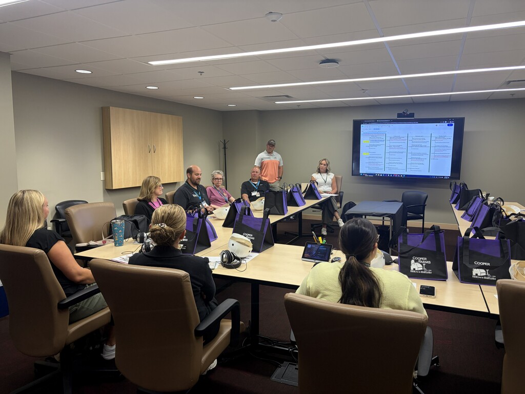 People in a conference room, sitting at tables with laptops and facing a large screen.