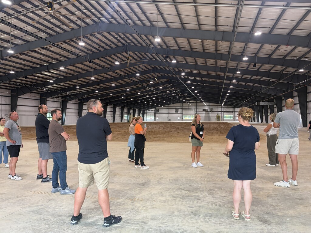 A group of people stands in a large, open, metal-roofed warehouse with a concrete floor.